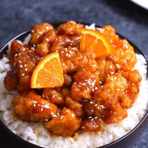 A serving of Chinese Orange Chicken garnished with sesame seeds and orange wedges on a bed of steamed rice