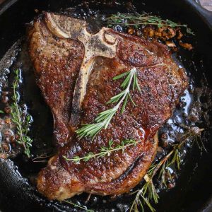 Overhead view of searing a t-bone steak in a very hot cast iron frying pan with the fat sizzling around the meat